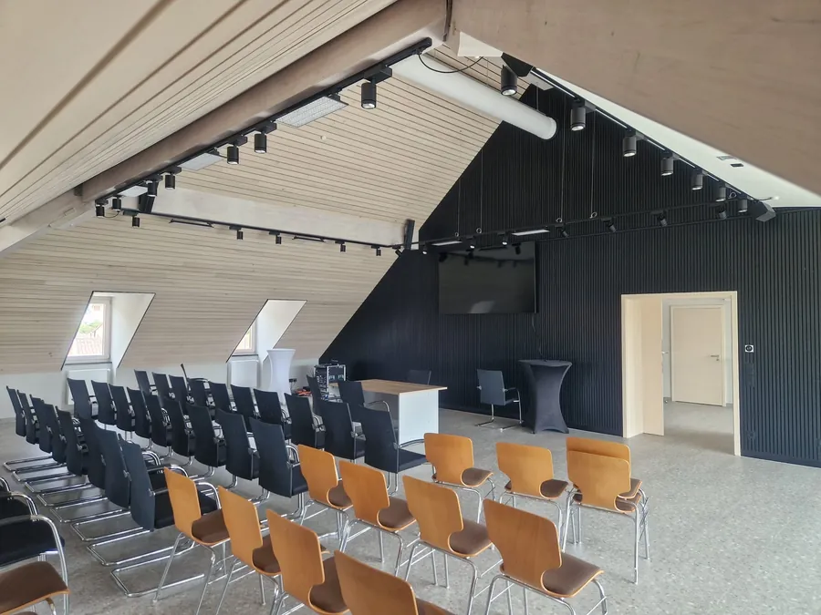 Bestuhlter Saal mit heller Holzdecke, dunkler Akustikwand und Strahlern auf Stromschienen
